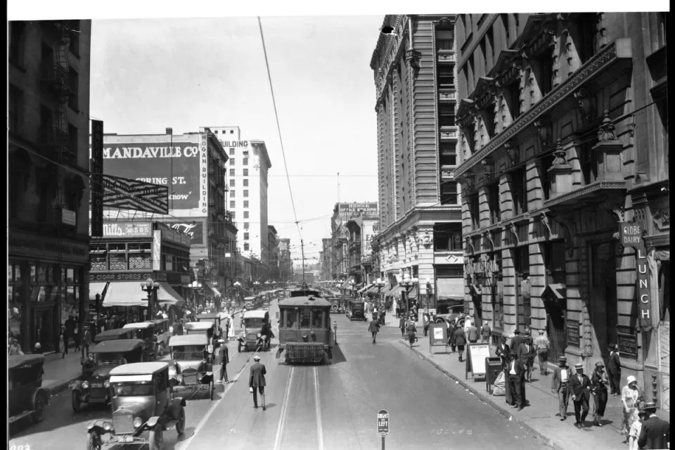 Black-and-white 1924 photo of a street in Los Angeles with a street car, cars driving in the other lane over street car tracks and pedestrians walking on sidewalk and middle of street.